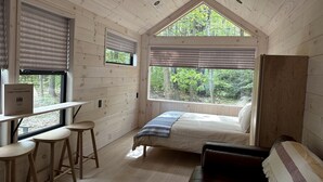 A cabin interior with a bed, a desk with stools, a window with blinds, and a view of the forest.