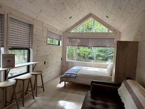 A cabin interior with a bed, a desk with stools, a window with blinds, and a view of the forest. - Nordic-Inspired Tiny Cabin Near Lake George & Hudson River, New York (Corinth)