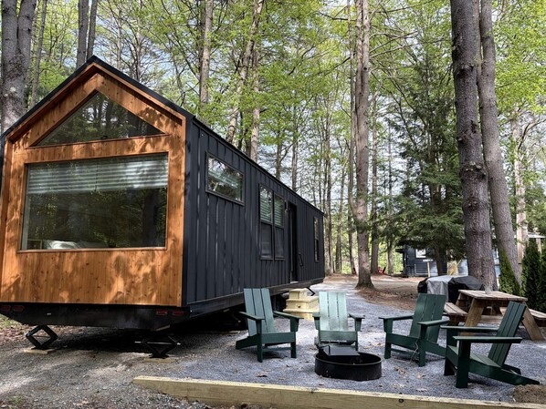 Una cabaña pequeña con fachada de madera y revestimiento negro, rodeada de árboles. Hay sillas Adirondack verdes y una mesa de picnic afuera.