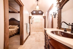 A narrow hallway leading to a bedroom with a bed, a bathroom with a sink and mirror, and a chandelier.