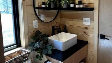 A wooden cabin bathroom with a white sink, a mirror, a shelf with plants, and a window.