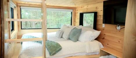 A wooden cabin bedroom with a bed, pillows, a TV, and large windows.