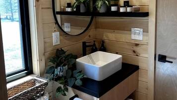 A wooden cabin bathroom with a white sink, a mirror, a shelf with plants, and a window.