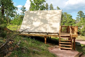 Una terraza de madera con un toldo, rodeada de árboles y terreno natural.