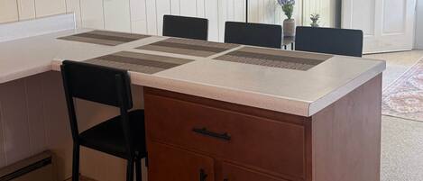 Un coin repas avec une table en bois, des chaises, un téléviseur fixé au mur et une porte visible au fond.