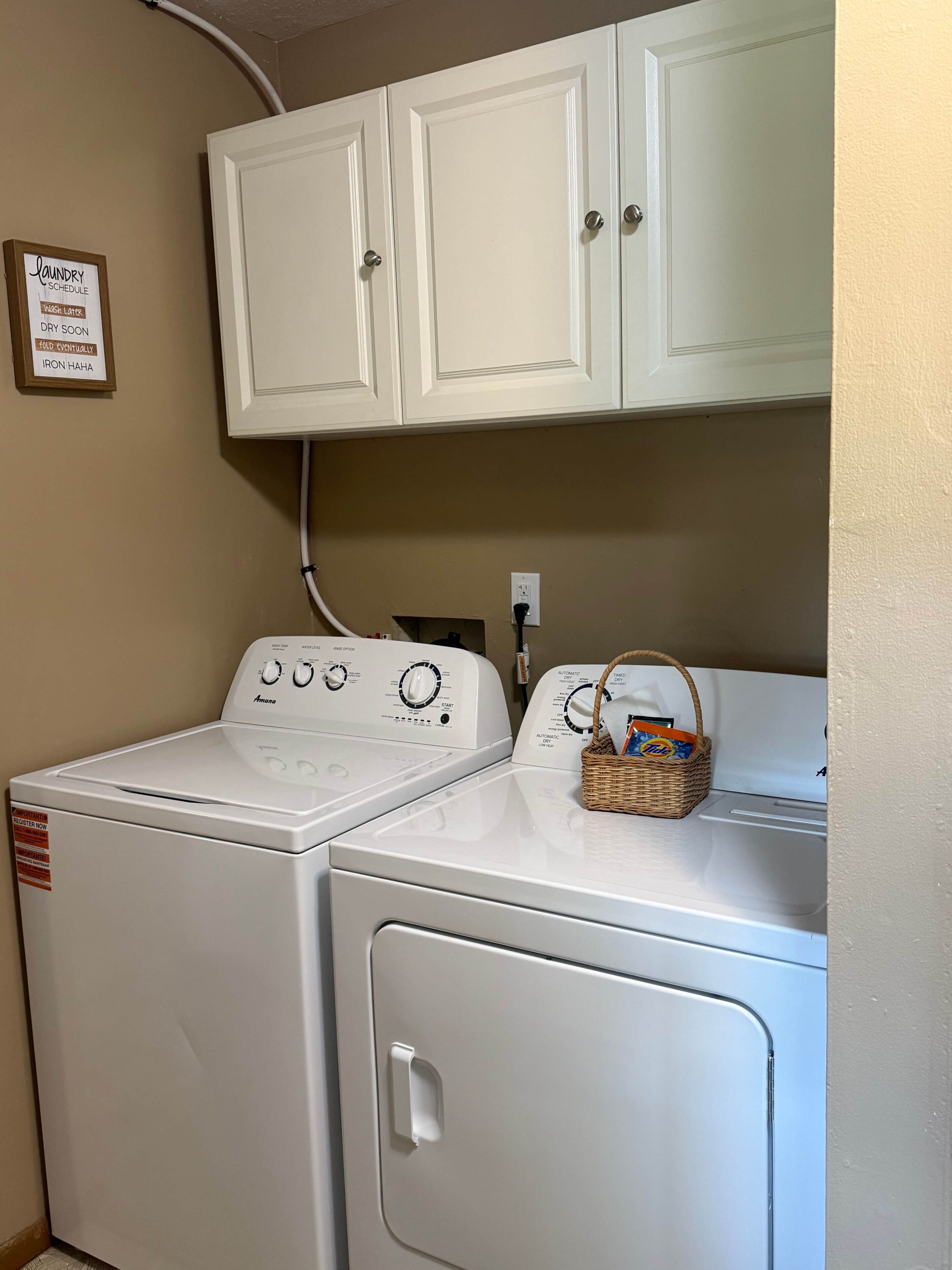 Une buanderie avec deux machines à laver et deux sèche-linge blancs, un panier en osier posé sur le sèche-linge et un tableau indiquant les jours de lessive accroché au mur.