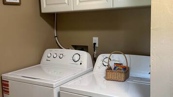 Une buanderie avec deux machines à laver et deux sèche-linge blancs, un panier en osier posé sur le sèche-linge et un tableau indiquant les jours de lessive accroché au mur.