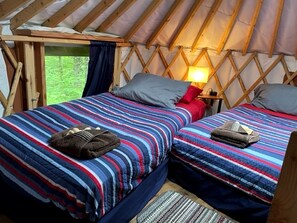A cozy interior of a yurt with two beds, each with a striped blanket and pillows, a small table with a lamp, and a view of greenery outside.