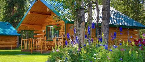 A log cabin with a porch, surrounded by a garden with various flowers and trees.