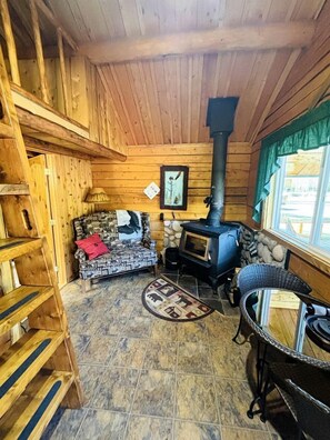 A wooden cabin interior with a stone fireplace, a patterned sofa, a dining table, and a window with a view.