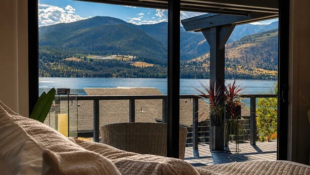 A room with a large window overlooking a lake and mountains, a balcony with outdoor furniture, and a view of the landscape. - Designer Lakefront Condo E3, Wapato Point Chelan (Manson)
