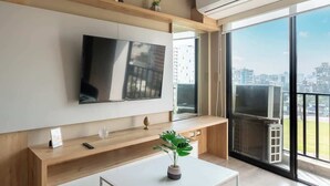 Una sala de estar moderna con televisor de pantalla plana, muebles de madera, una planta pequeña y un balcón con vista a la ciudad.