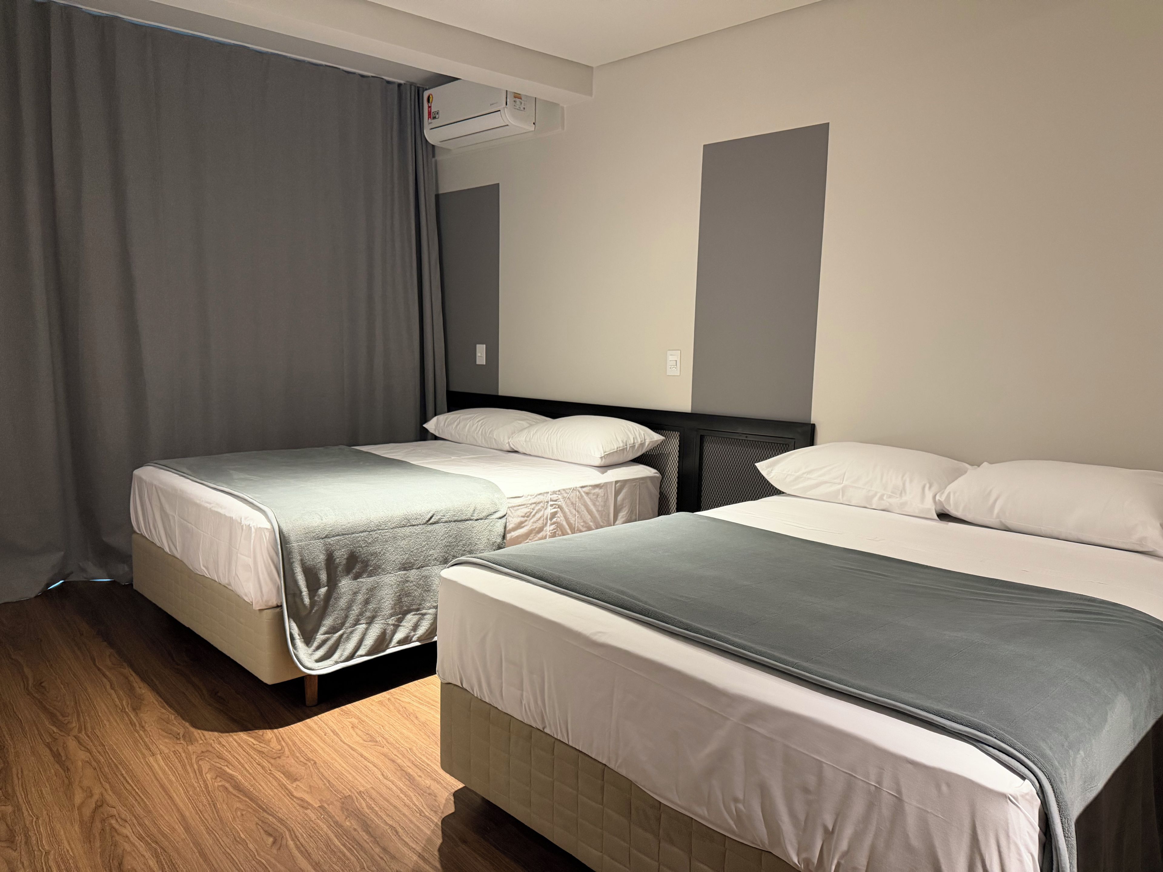 Habitación de hotel con dos camas, una cortina gris y un equipo de aire acondicionado en la pared.