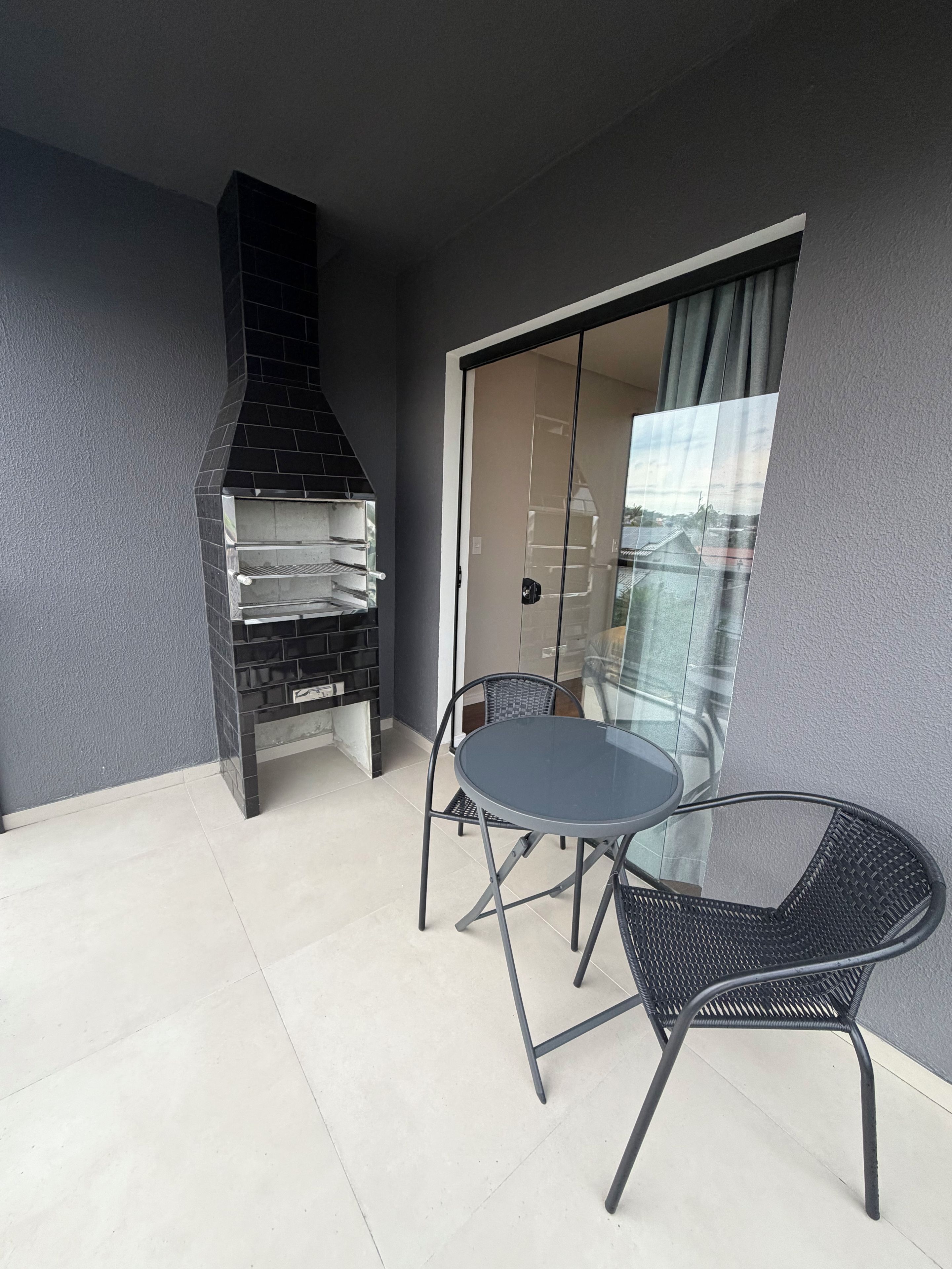 Ein Balkon mit einem schwarzen Grill, einer Glastür, die zu einem Raum führt, und einem runden Tisch mit Stühlen.