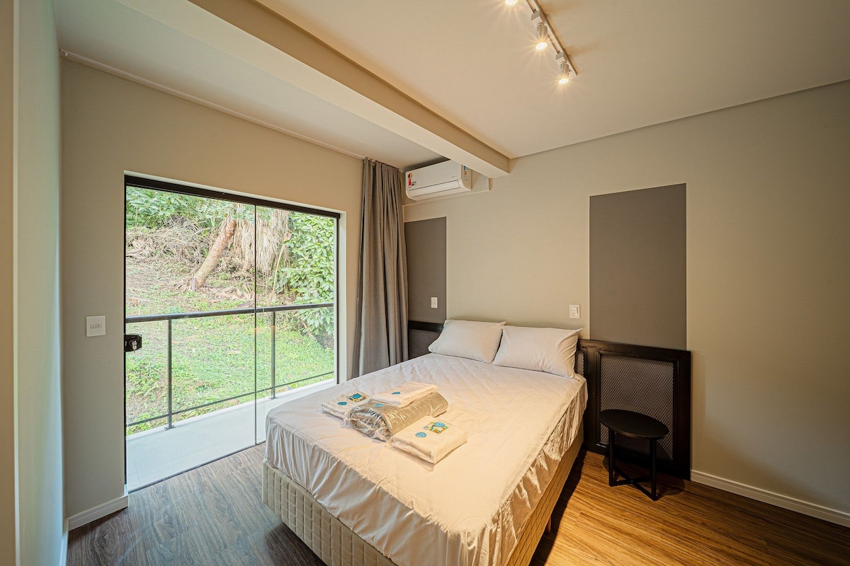 A bedroom with a large bed, a small table, a window with a view of greenery, and an air conditioning unit.