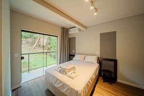 A bedroom with a large bed, a small table, a window with a view of greenery, and an air conditioning unit. - Apez Smart Studio (Joinville)