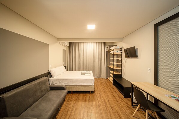 A compact hotel room with a bed, a sofa, a desk, and a TV. - Apez Smart Studio (Joinville)