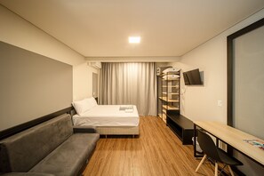 A compact hotel room with a bed, a sofa, a desk, and a TV. - Apez Smart Studio (Joinville)