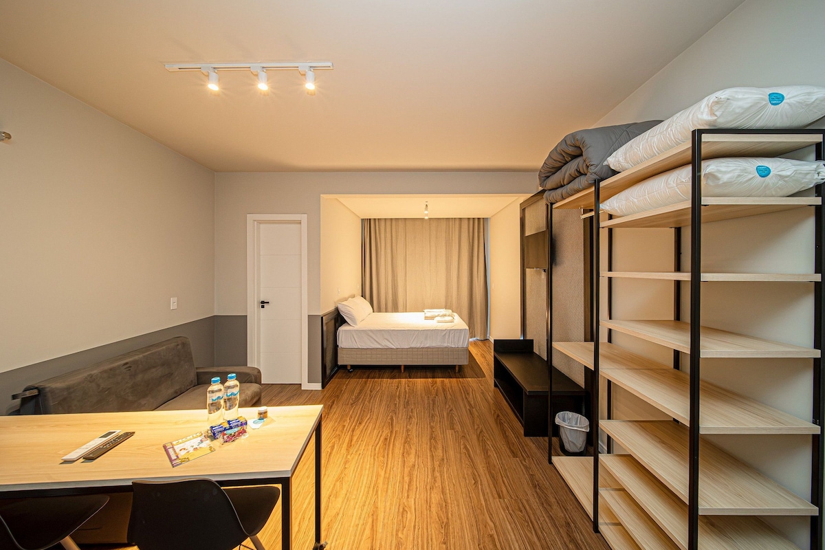 A compact hotel room with a bunk bed, a single bed, a small table with a chair, and a desk with a chair. There is a door and a window with curtains.