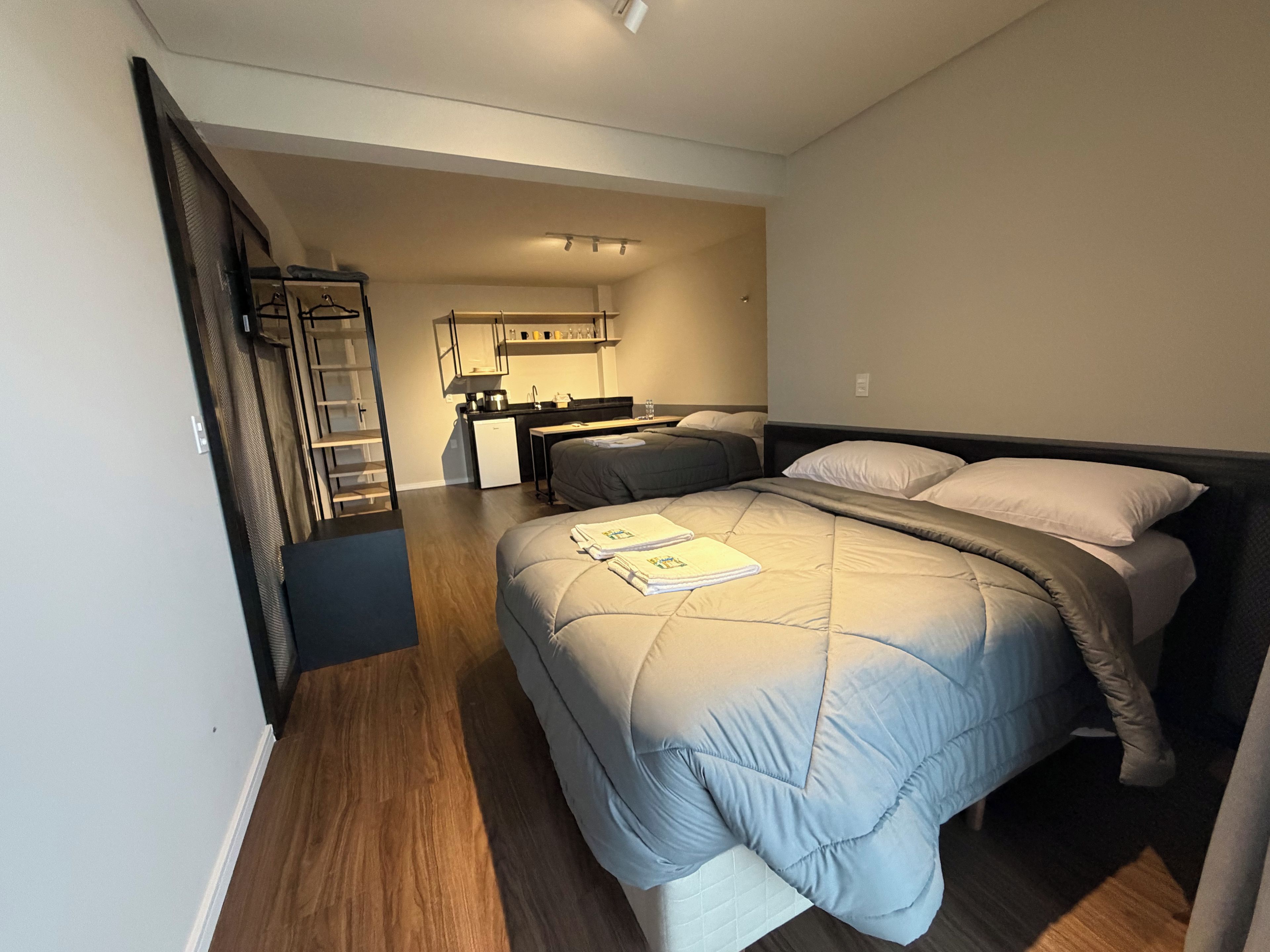 Un dormitorio con cama, mesitas de noche, un armario y una kitchenette al fondo.