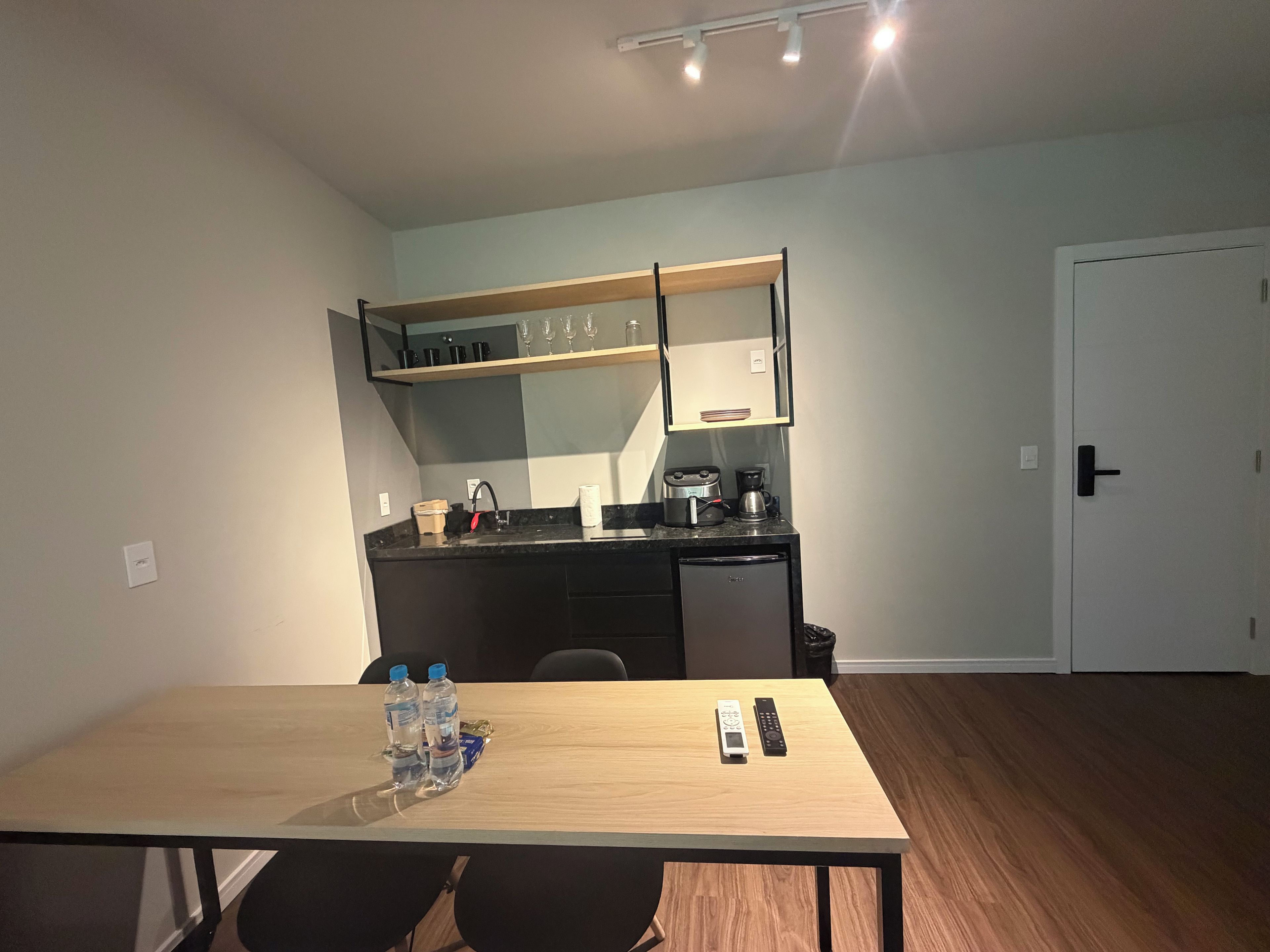Cocina compacta con encimera, fregadero y cafetera. Hay una mesa con dos botellas de agua y un control remoto.