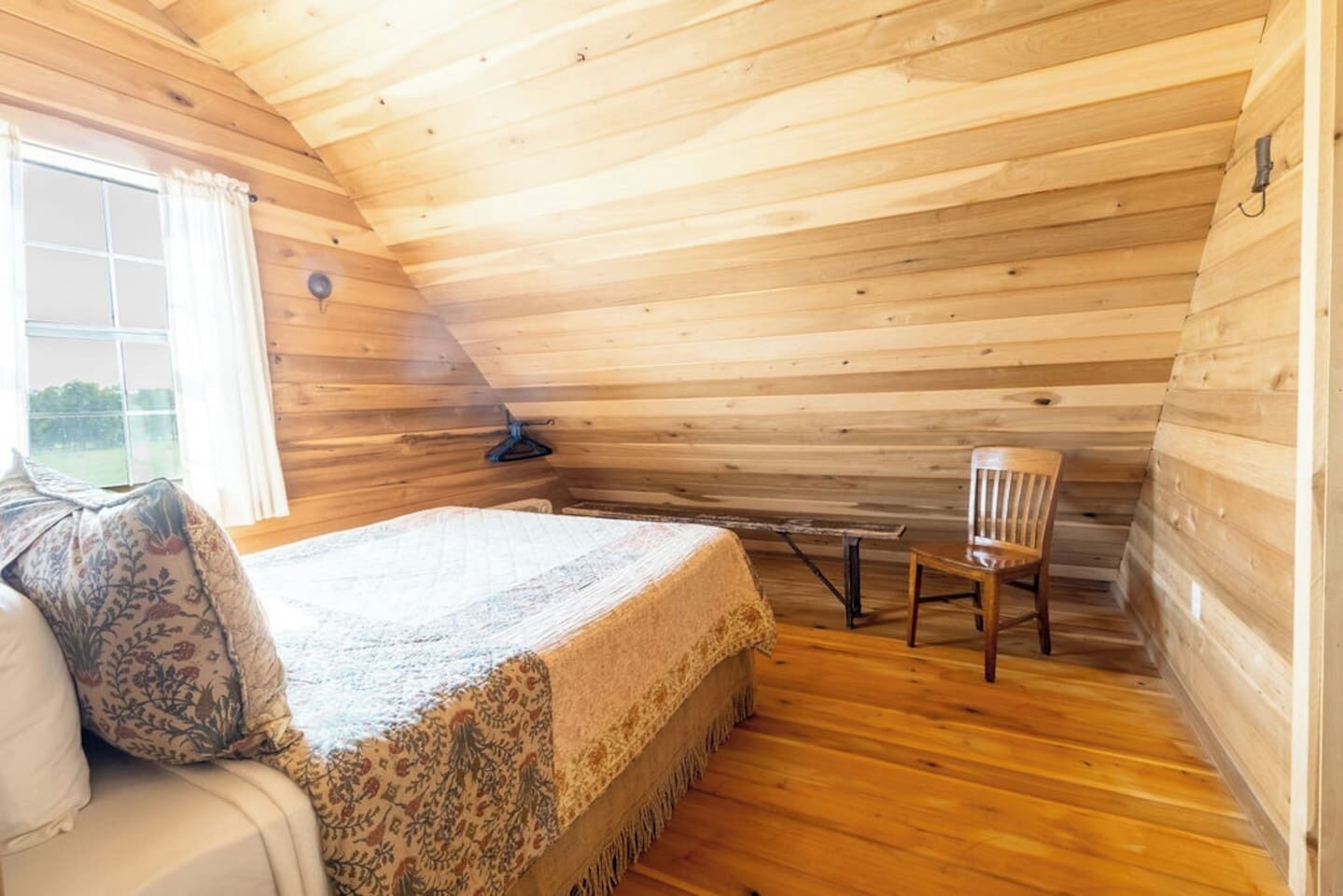 Een knus kamer in een blokhut met een bed, een stoel, een raam en een houten plafond.