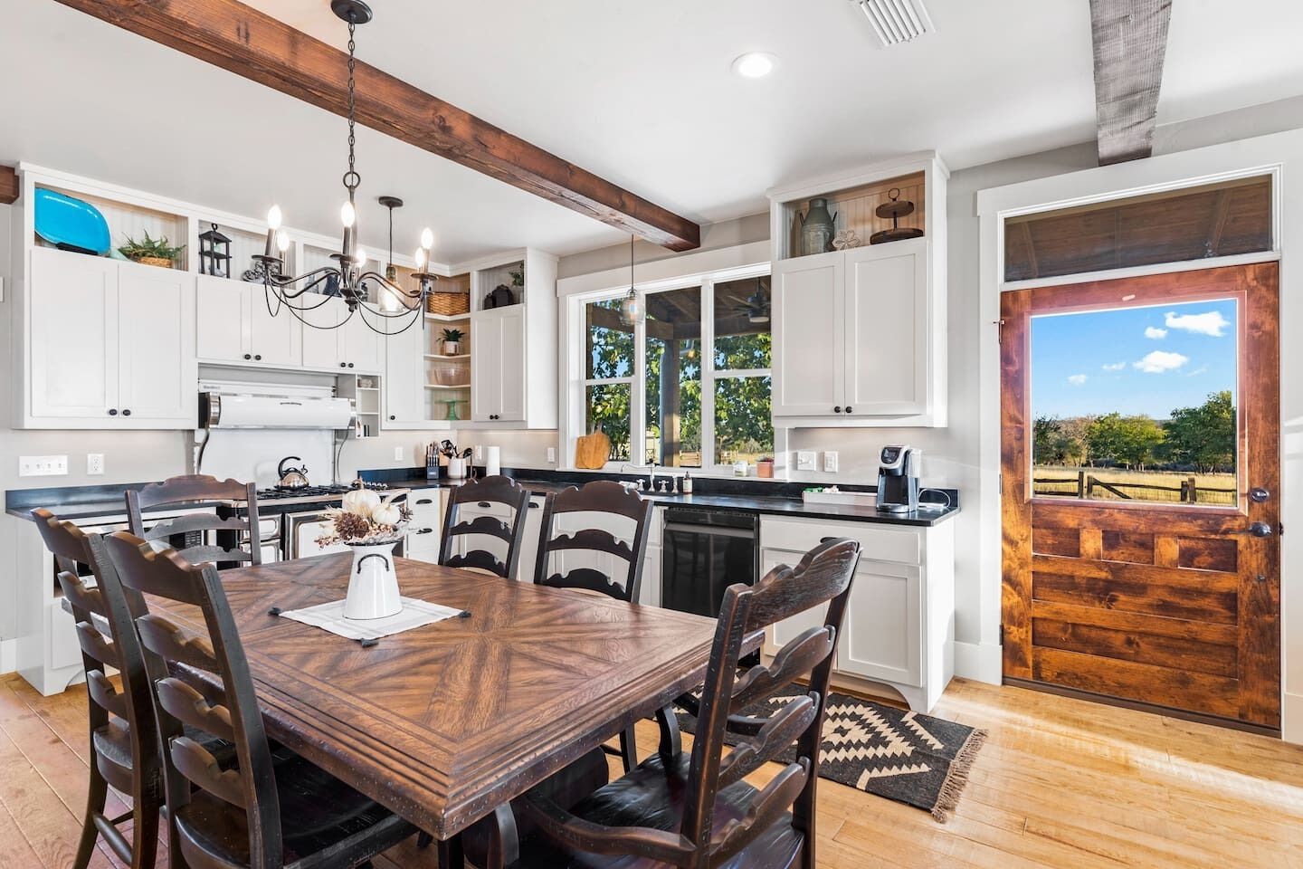 A modern kitchen with white cabinets, a wooden dining table, and a view of a field through a door.