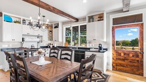 A modern kitchen with white cabinets, a wooden dining table, and a view of a field through a door. - TimberRose Ranch | Hot Tub + Hill Country Views (Fredericksburg)