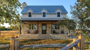 A two-story stone house with a covered porch, a metal roof, and a fenced yard. - TimberRose Ranch | Hot Tub + Hill Country Views (Fredericksburg)