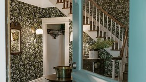 Eine Treppe mit floralem Tapetenmuster, ein runder Tisch mit einem Metalltopf und eine Tür, die in einen Raum mit weißer Wand und Spiegel führt.