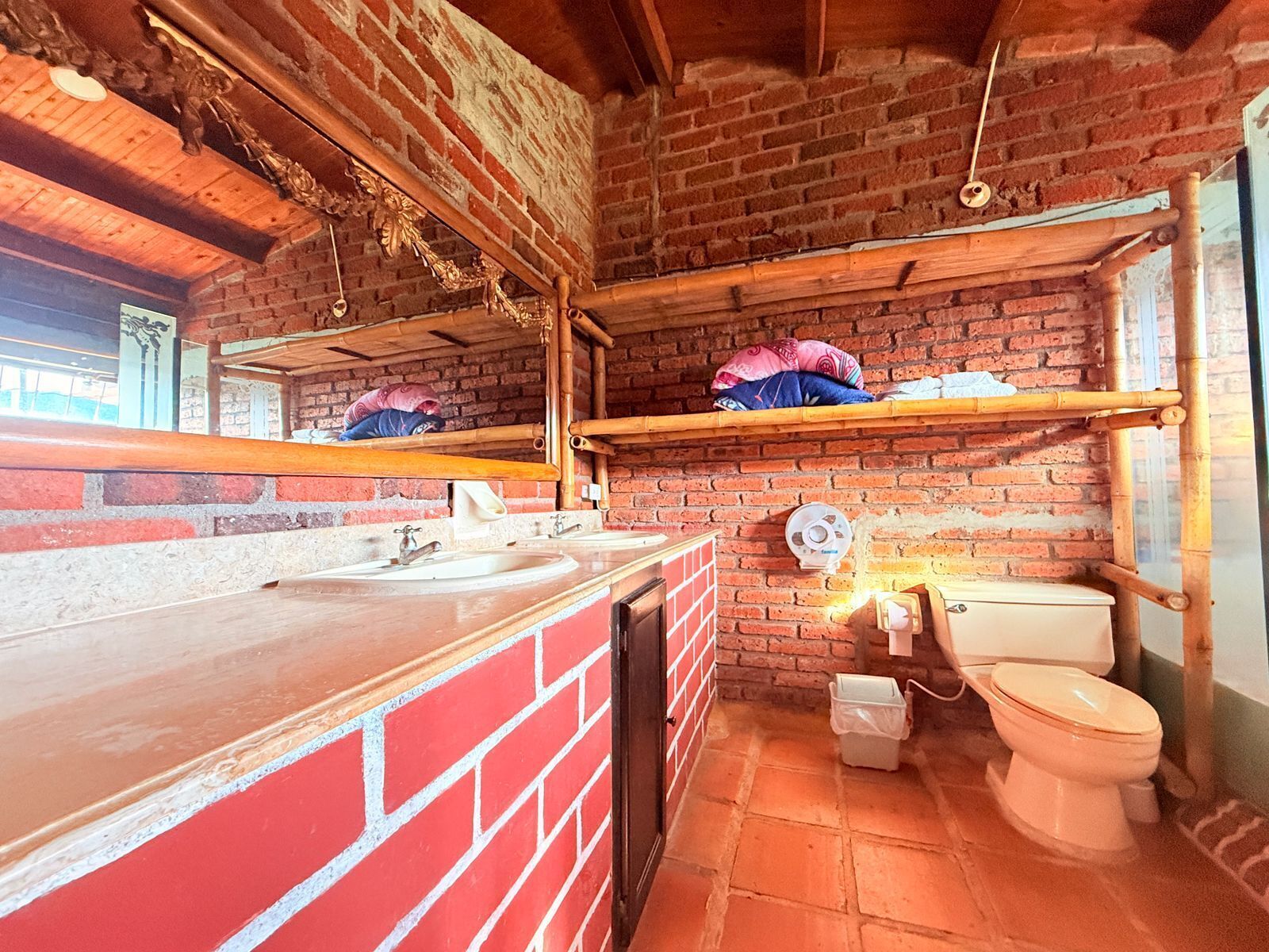 Een badkamer met een bakstenen muur, houten balken, een wastafel, toilet en een spiegel.