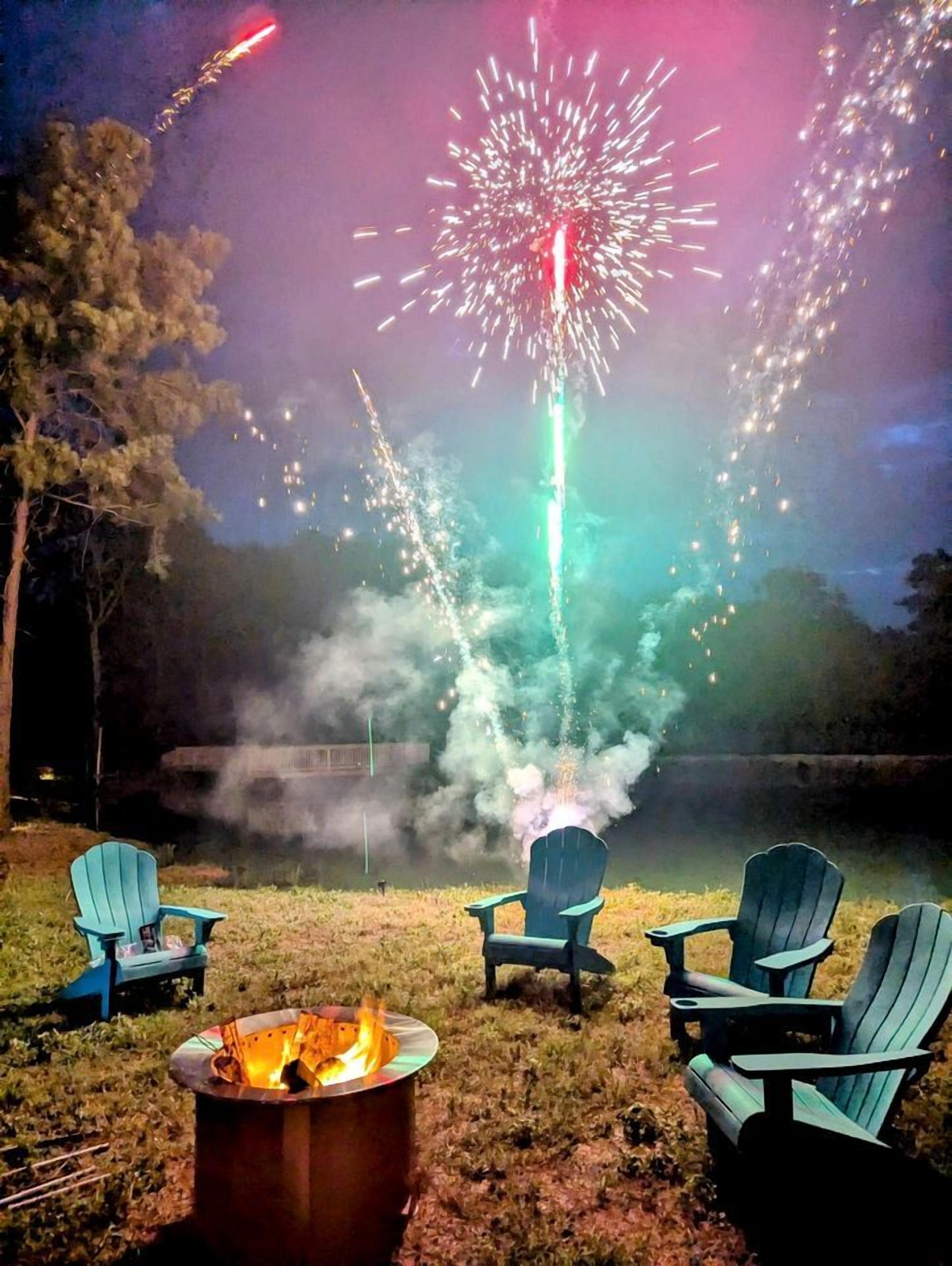 Escena nocturna al aire libre con un brasero, tres sillas Adirondack y un espectáculo de fuegos artificiales al fondo.