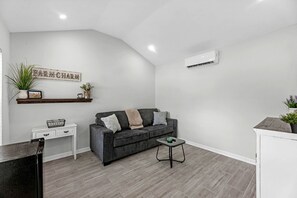 Een moderne woonkamer met een grijze bank, een klein bijzettafeltje, een witte dressoir, een potplant en een wandairconditioner.