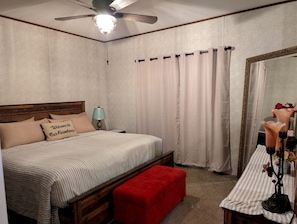 Een slaapkamer met een bed, nachtkastje, rode bank, plafondventilator, spiegel en gordijnen.