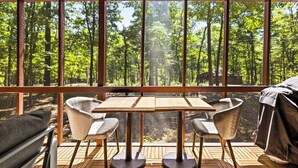 A room with a large window overlooking a forest, a wooden table with chairs, and a covered grill.