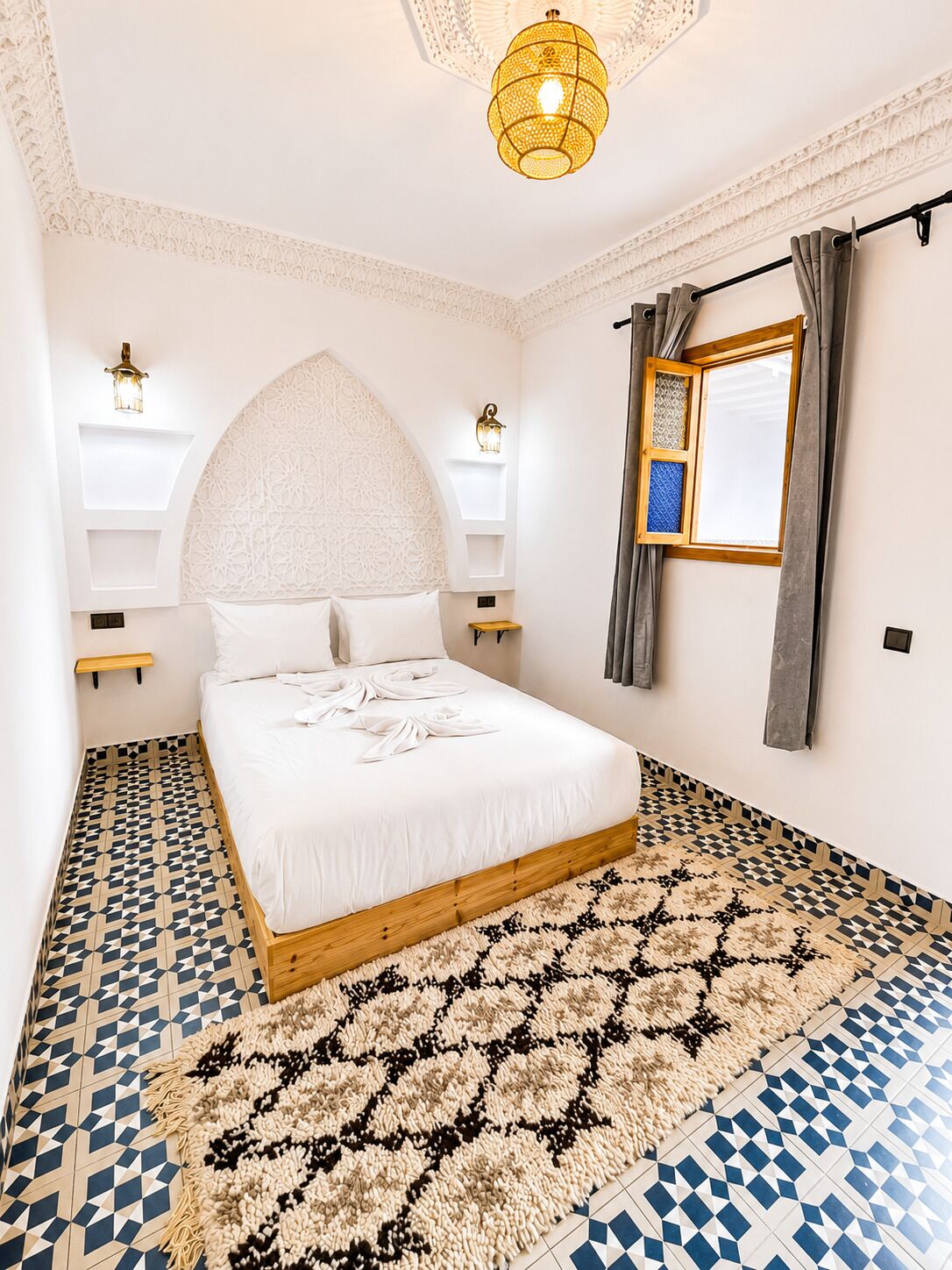 A bedroom with a bed, a large window with curtains, a decorative ceiling light, and patterned flooring.