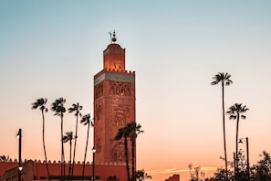 A tall tower with intricate architectural details and a clock at the top, surrounded by palm trees against a sunset sky. - Riad Le Sultan Rouge And Spa (Marrakech)