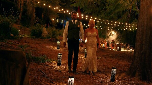 Una pareja caminando por un sendero bordeado de luces colgantes, entre árboles.