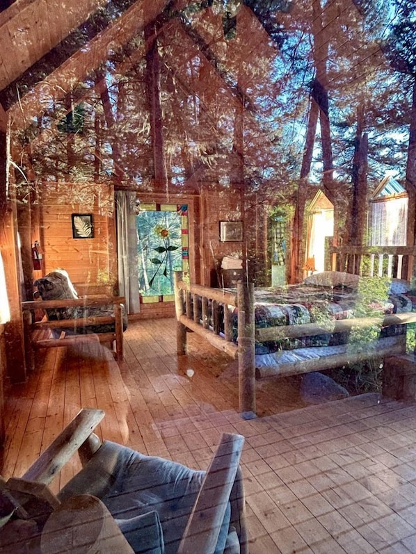 Interior rústico de una cabaña con pisos de madera, una cama con dosel y un ventanal con vistas a los árboles.