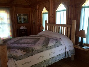 A wooden cabin room with a bed, a nightstand, a lamp, a framed picture on the wall, and a window with curtains. - Romantic Cabin Rental for Couples in Secluded Woodlands near Butte, Montana (Hall)