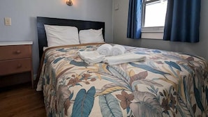 A bedroom with a floral bedspread, a nightstand, a wall-mounted light, and a window with curtains. - Beachfront Escape with Breezy Views in North Truro, Massachusetts (North Truro)