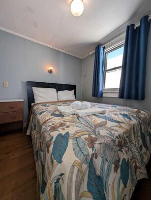 A bedroom with a floral bedspread, a nightstand, a wall-mounted light, and a window with curtains. - Beachfront Escape with Breezy Views in North Truro, Massachusetts (North Truro)