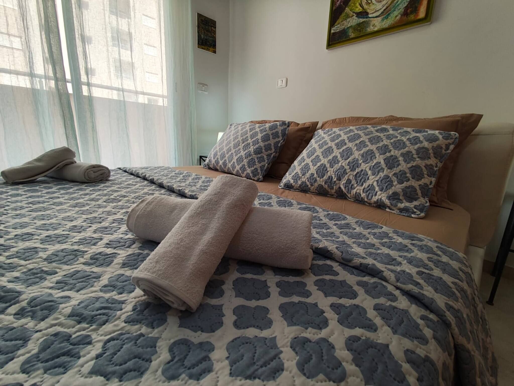 A neatly made bed with a patterned bedspread, two pillows, and rolled towels on a hotel room.