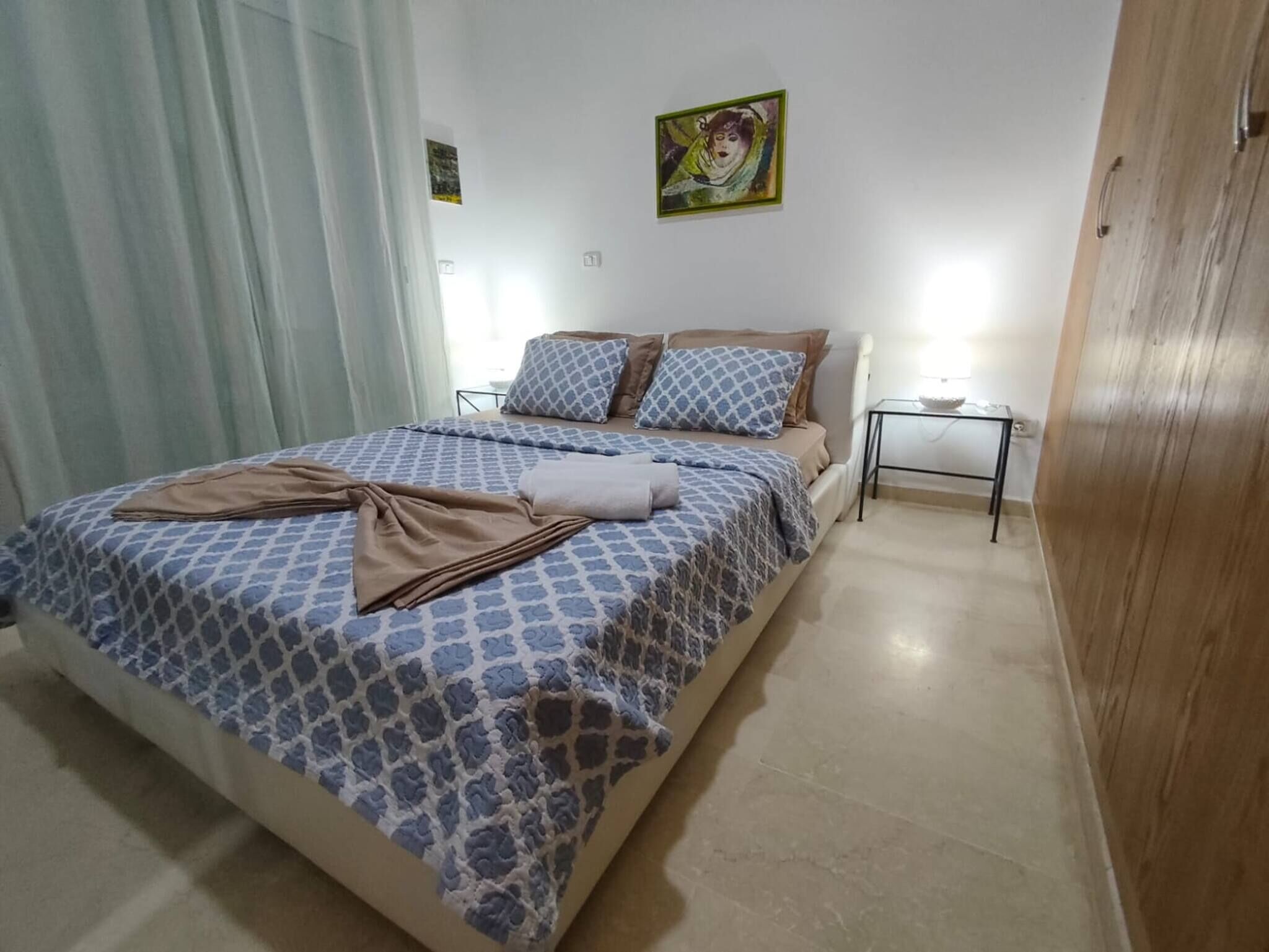 Een slaapkamer met een bed, een nachtkastje, een lamp en een schilderij aan de muur.