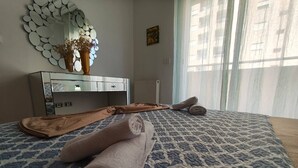 Une chambre à coucher avec une couvre-lit à motifs, une table de chevet ornée d’un vase et de fleurs, un miroir à cadre circulaire et une fenêtre avec des rideaux.