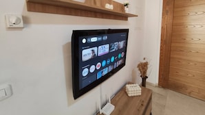 Un téléviseur à écran plat fixé au mur, une étagère en bois au-dessus, et une petite table sur laquelle est posé un objet décoratif.