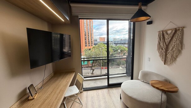 Quarto moderno com televisão de tela plana, uma cadeira branca arredondada, uma pequena mesa de madeira e uma janela grande com vista para a cidade.
