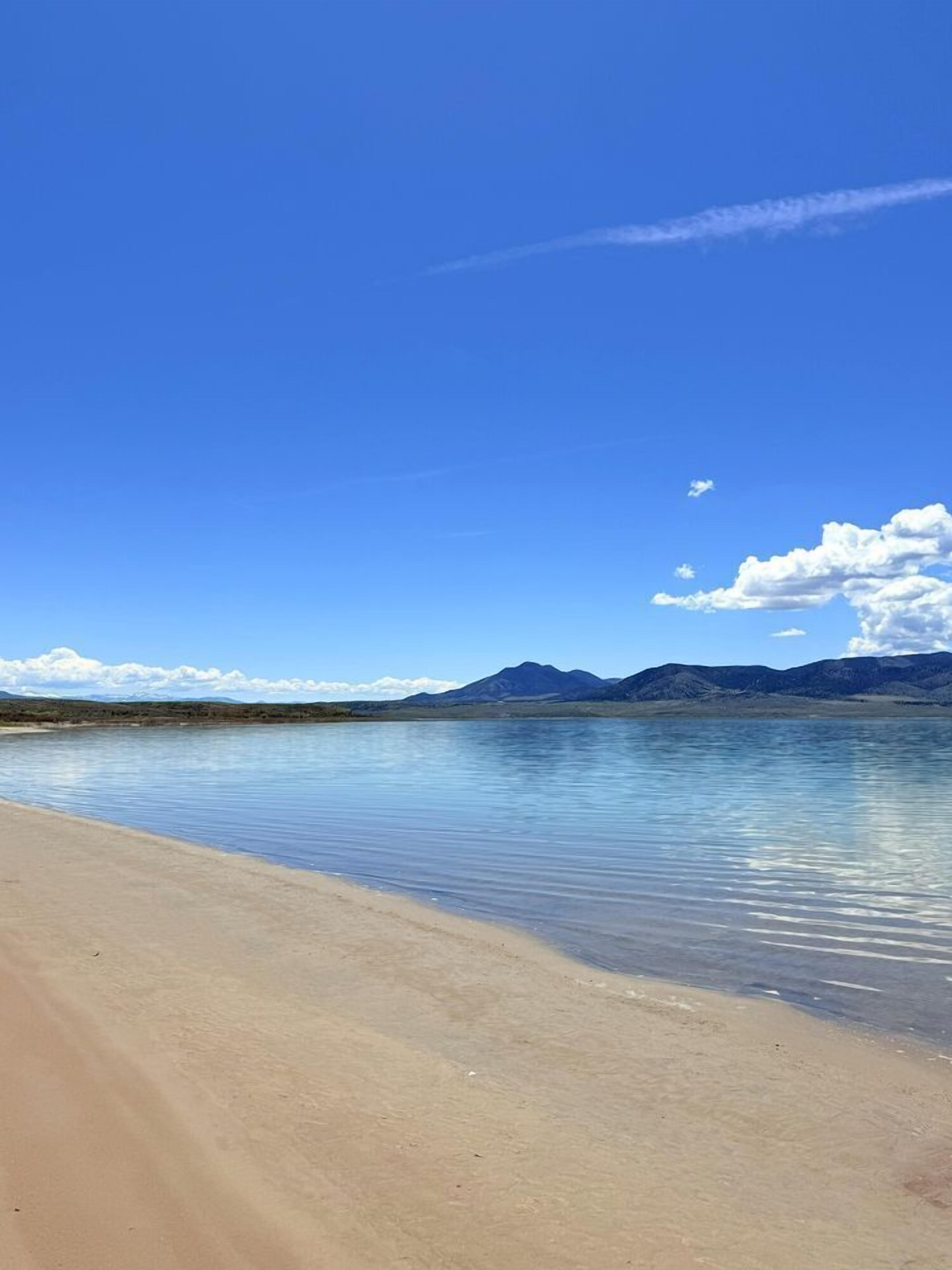 A beach with clear water, a sandy shore, and distant mountains under a blue sky.