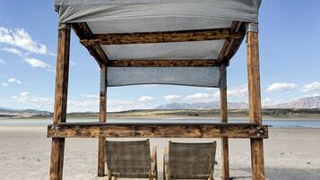 A wooden beach chair with a grey canopy, two wicker chairs, and a view of a lake and mountains.