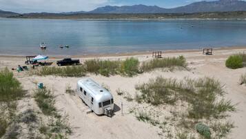 A campsite with a trailer, a truck, and a boat on a lake with mountains in the background.